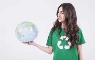 Sustentabilidade na prática mpresa - Uma jovem de pele branca, sorri segurando um globo do mundo, ela usa camiseta verde e símbolo branco de reciclagem.